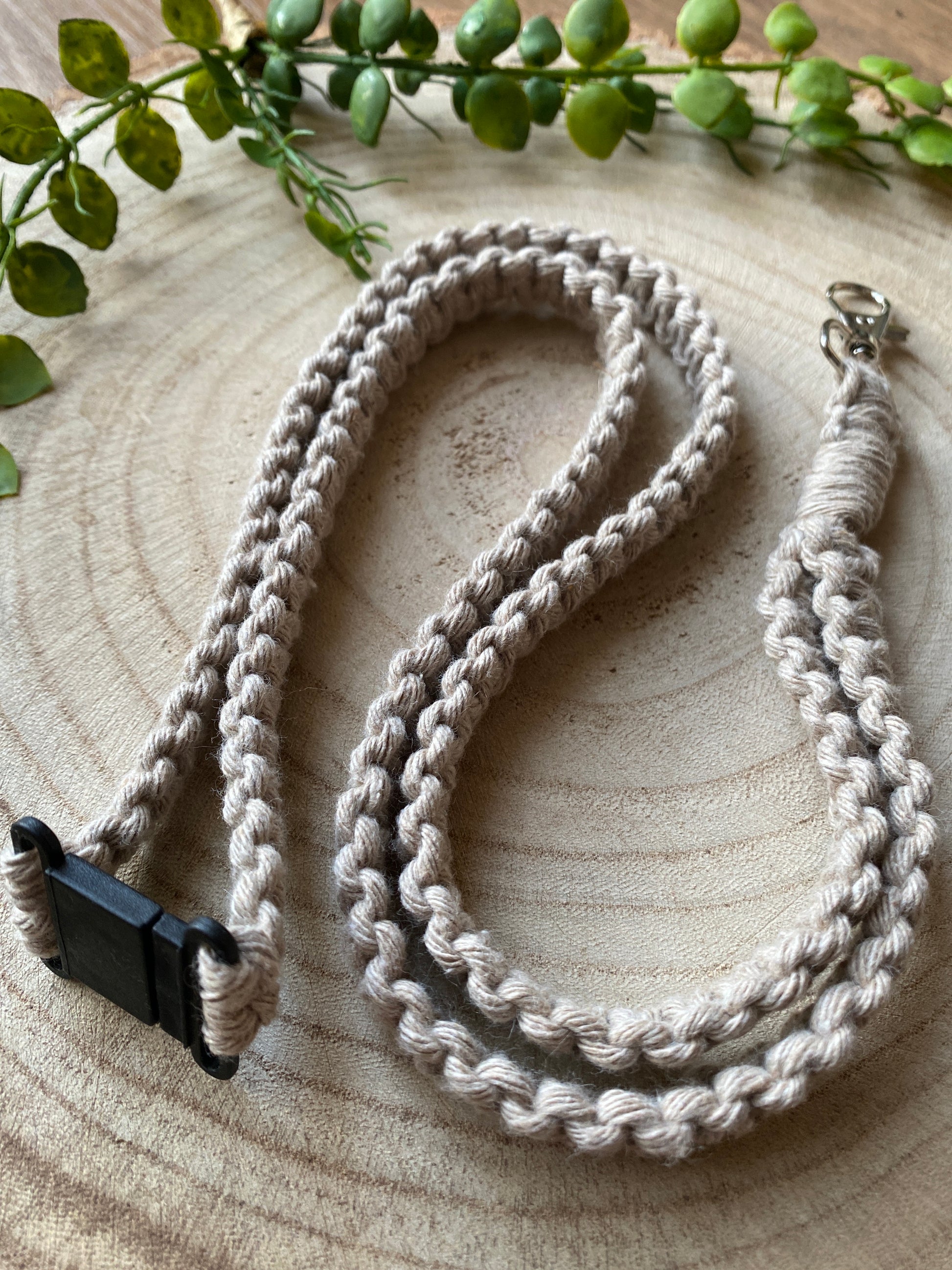 A handmade, braided beige lanyard with a black plastic breakaway clasp and a silver metal clasp is laid out in a loose loop on a wooden surface. Greenery surrounds the *Macra-Made-With-Love* Eco friendly lanyard badge holder - 34”, providing a natural backdrop.