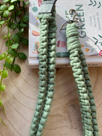 A close-up view of a green, Macra-Made-With-Love Eco friendly lanyard badge holder - 34” placed on a wooden surface. The intricately braided, handmade lanyard is attached to a silver clip designed as an ID badge holder. A book titled "LIVE GREEN" with plant illustrations is partially visible underneath, next to some green plant leaves.