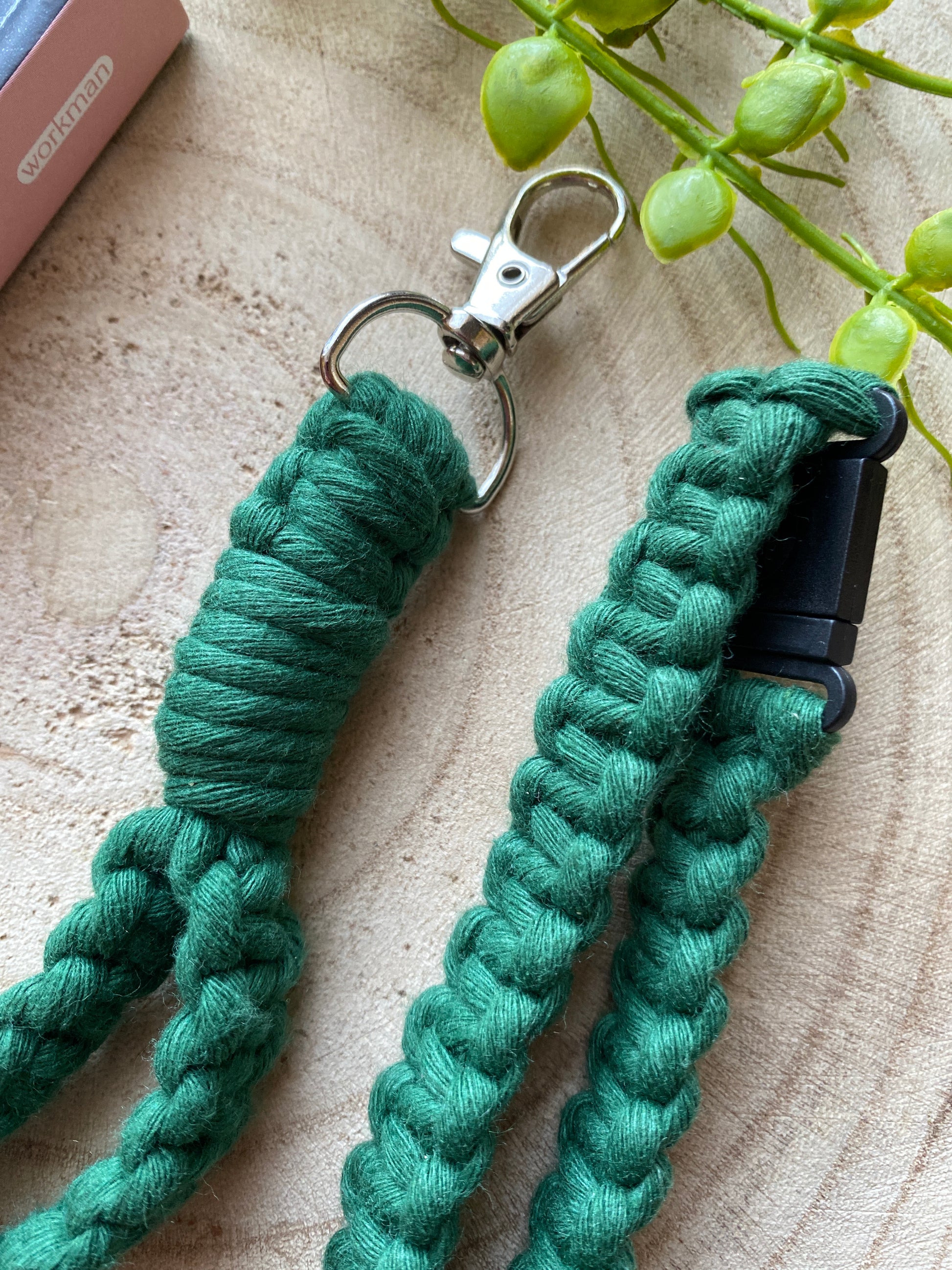 Close-up of an eco-friendly, green, braided, rope-like Eco friendly lanyard badge holder - 34” from Macra-Made-With-Love with a metal clasp on a wooden surface. The handmade lanyard is intricately woven, showcasing detailed craftsmanship. There's a small black plastic connector on one end and a partial view of a green plant in the background.