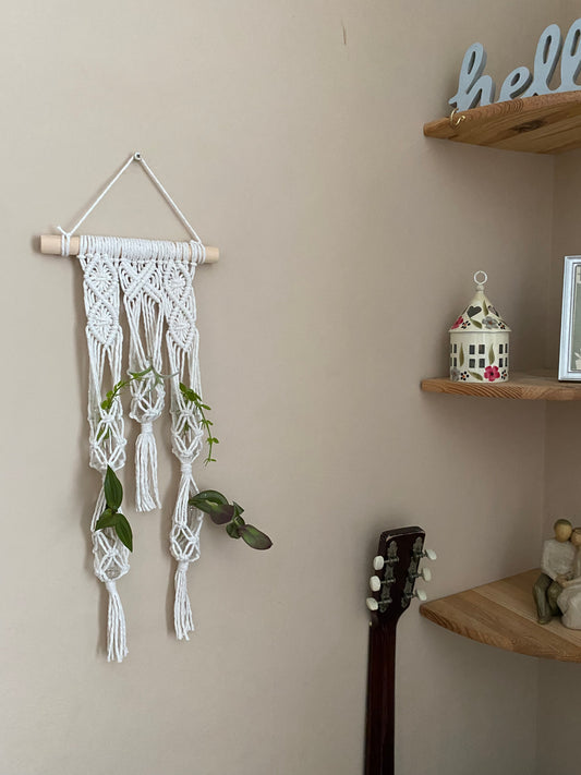 A cozy corner features a Macra-Made-With-Love Macrame wall hanger houseplant propagation station holding small green plants, perfect for houseplant propagation. A guitar rests against the wall. Wooden shelves hold a lantern, small decorative house, framed picture, and a "hello" sign. The beige wall adds warmth to this eco-friendly home décor space.