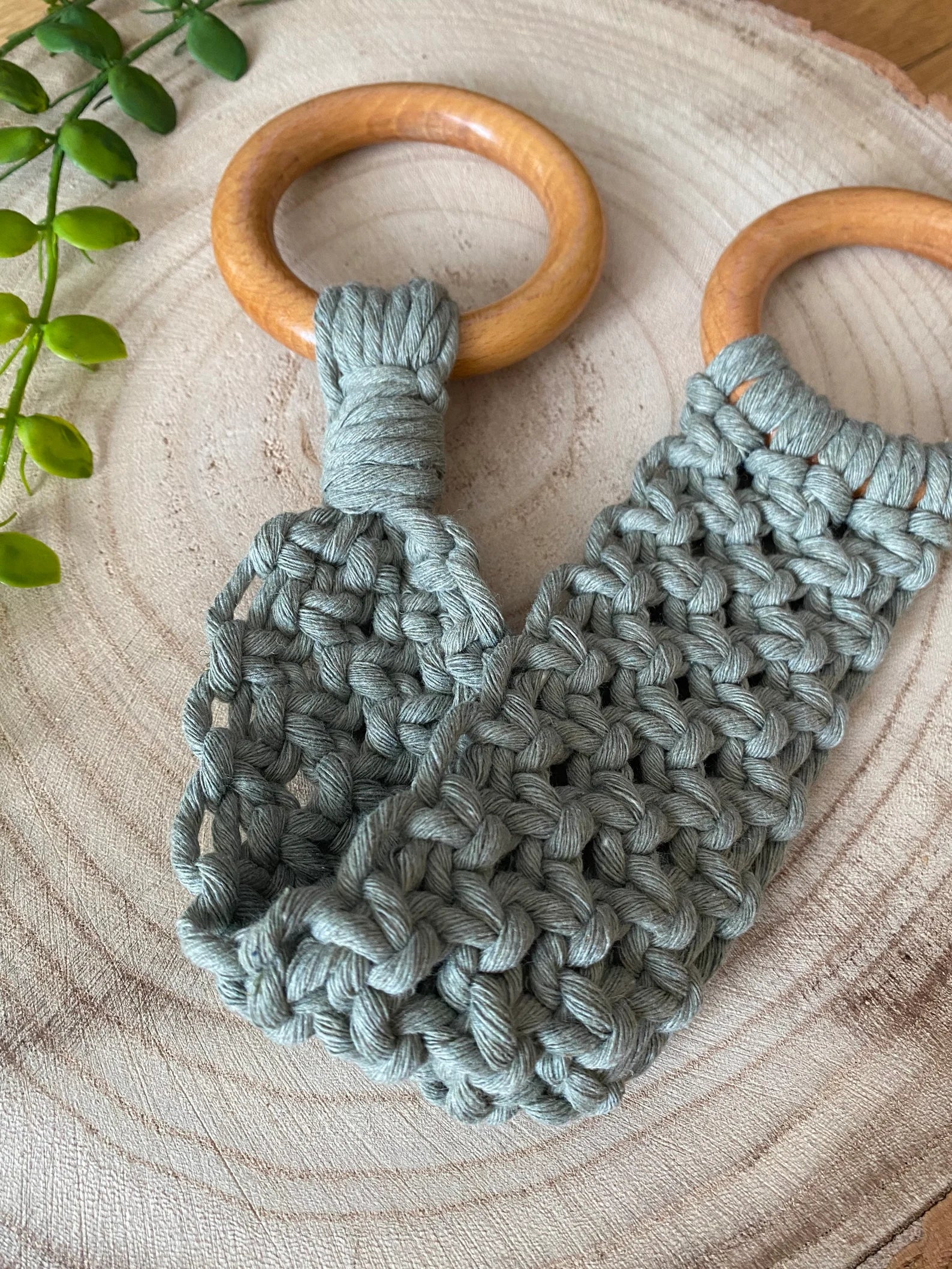 A handmade macrame knotted kitchen towel holder made with sage green cotton yarn and two large wooden rings, lays folded on a circular wooden board. 