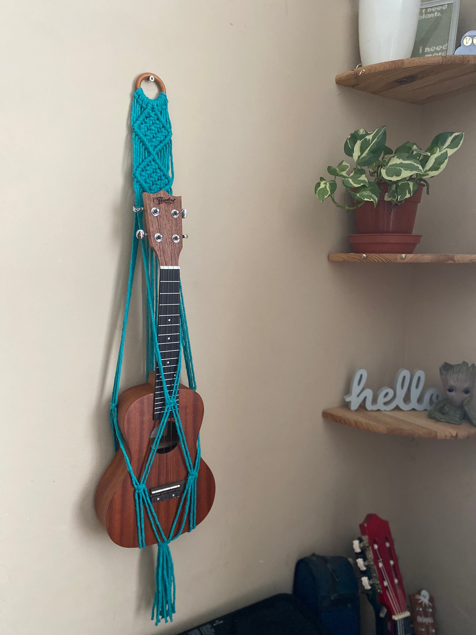 A dark brown ukulele sits inside of a handmade macrame wall hanging ukulele hanger, which is made with turquoise blue recycled cotton, and a repurposed wooden hanging ring. There are a set of corner shelves next to the hanger, with a small houseplant on. 