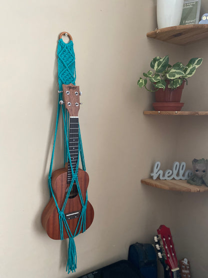 A dark brown ukulele sits inside of a handmade macrame wall hanging ukulele hanger, which is made with turquoise blue recycled cotton, and a repurposed wooden hanging ring. There are a set of corner shelves next to the hanger, with a small houseplant on. 
