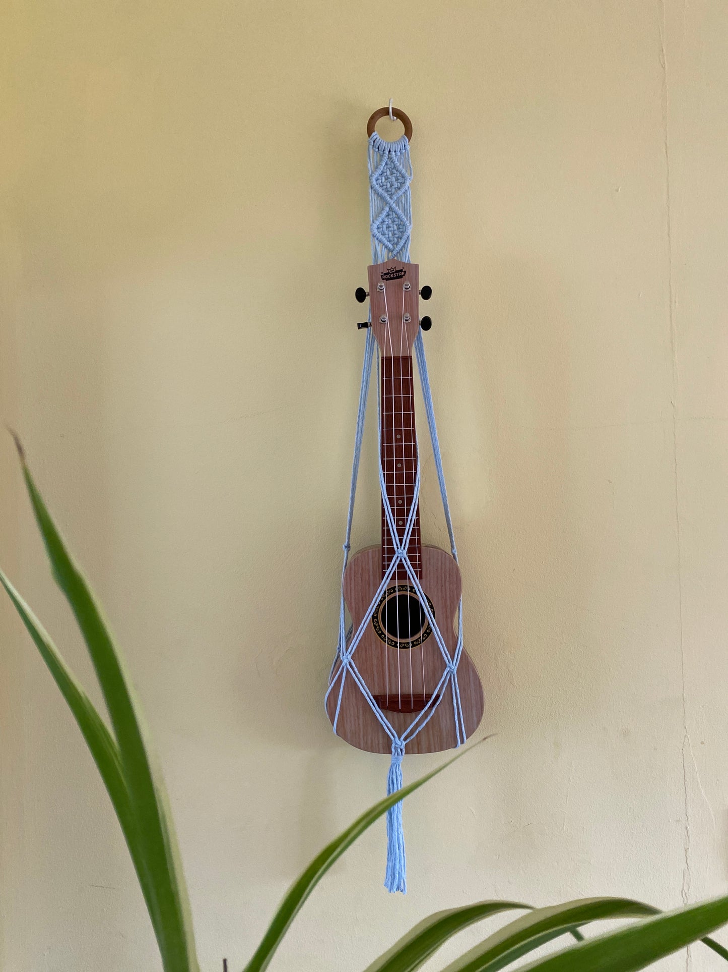 A light brown ukulele sits inside of a handmade macrame wall hanging ukulele hanger, which is made with light blue recycled cotton, and a repurposed wooden hanging ring. A large green leafy plant is at the front of the image. 