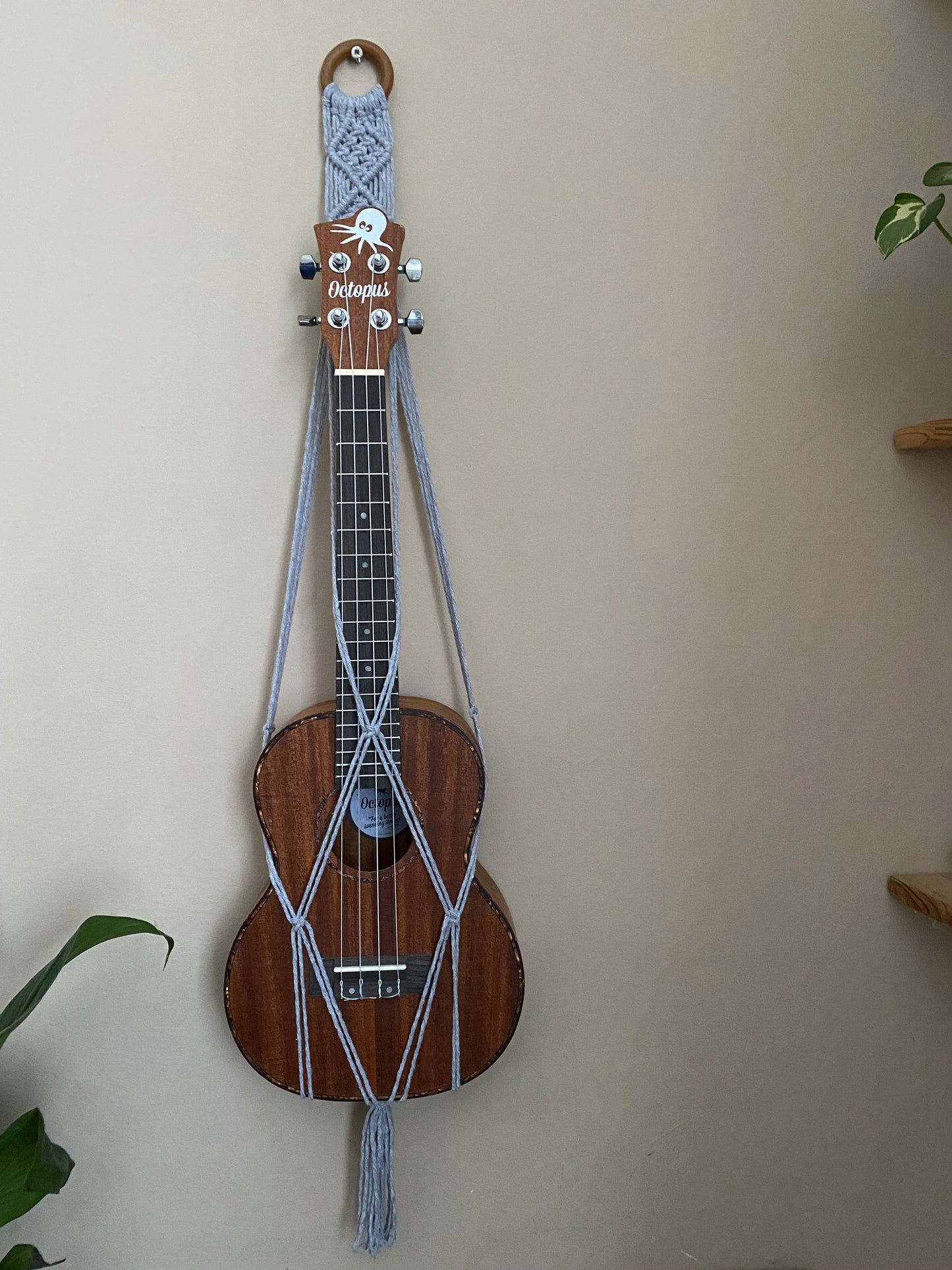 A dark brown ukulele sits inside of a handmade macrame wall hanging ukulele hanger, which is made with light grey recycled cotton, and a repurposed wooden hanging ring. 