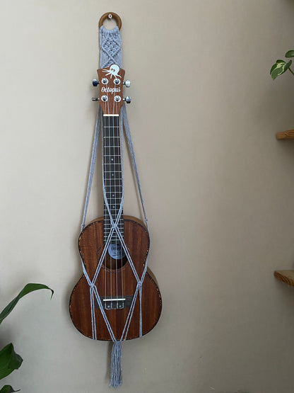 A dark brown ukulele sits inside of a handmade macrame wall hanging ukulele hanger, which is made with light grey recycled cotton, and a repurposed wooden hanging ring. 