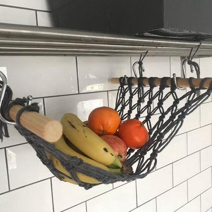 Bananas, oranges and apples hang in a macrame hammock made with dark grey recycled cotton and wooden dowels, underneath a metal shelf on a white tiled wall. 