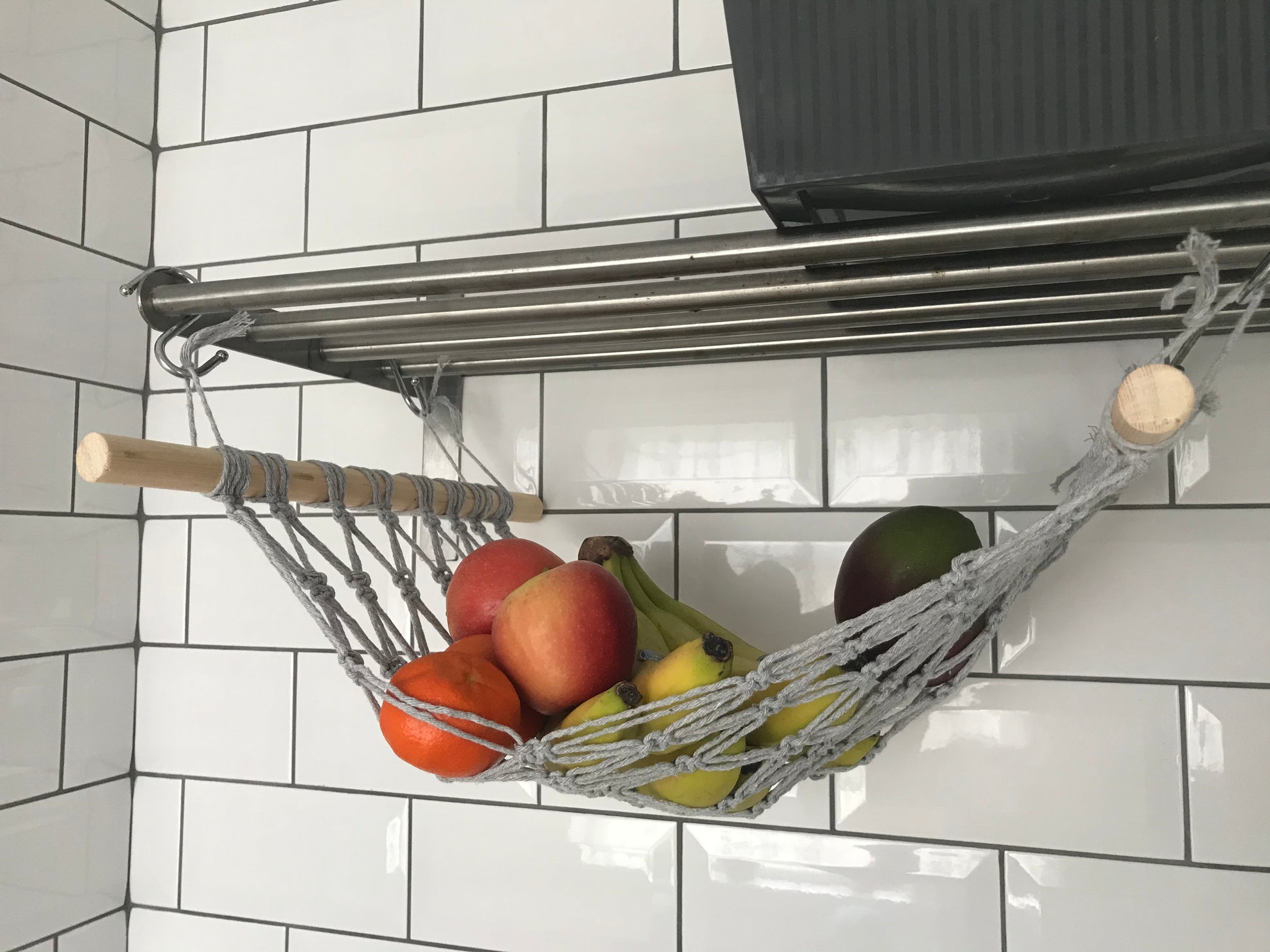 Various fruits hang in a macrame hammock made with light grey recycled cotton and wooden dowels, underneath a metal shelf, using metal S hooks. 