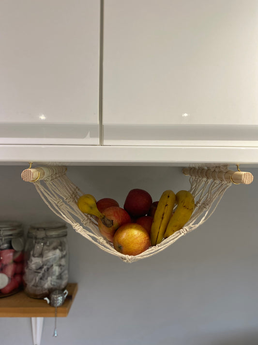 Bananas and apples hang in a macrame hammock made with natural recycled cotton and wooden dowels, underneath a white kitchen wall unit. There are glass jars with tea bags and coffee pods inside on a wooden shelf in the background. 