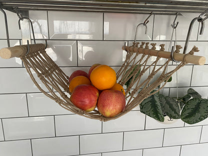 Oranges and apples hang in a macrame hammock made with taupe recycled cotton and wooden dowels, underneath a metal wall shelf using metal S hooks. 