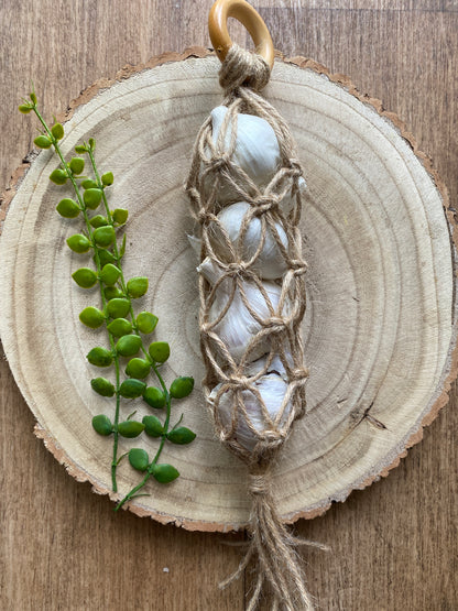 A handmade macrame garlic hanger made with jute twine and a wooden ring, laid on a wooden board with 4 fresh garlic bulbs inside. 