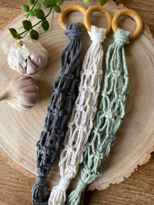 Three handmade macrame garlic hangers made with cotton yarn and wooden rings, laid across a round wooden board next to two fresh garlic bulbs. 