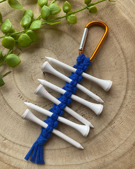 A knotted golf tee holder, made with blue yarn on an orange gold carabiner clip, laid on a wooden surface holding 6 white golf tees. 