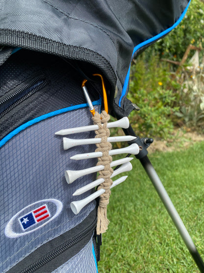 A knotted golf tee holder, made with beige yarn on an orange gold carabiner clip, attached to the side of a black golf bag, holding 6 white golf tees. 