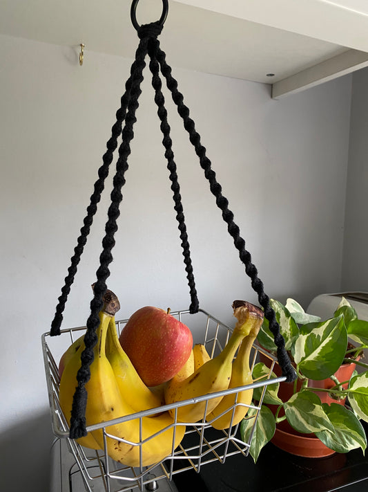Bananas and apples in a metal basket, suspended by black macrame ropes on a metal hoop, hanging under a white kitchen cabinet next to a variegated houseplant. 