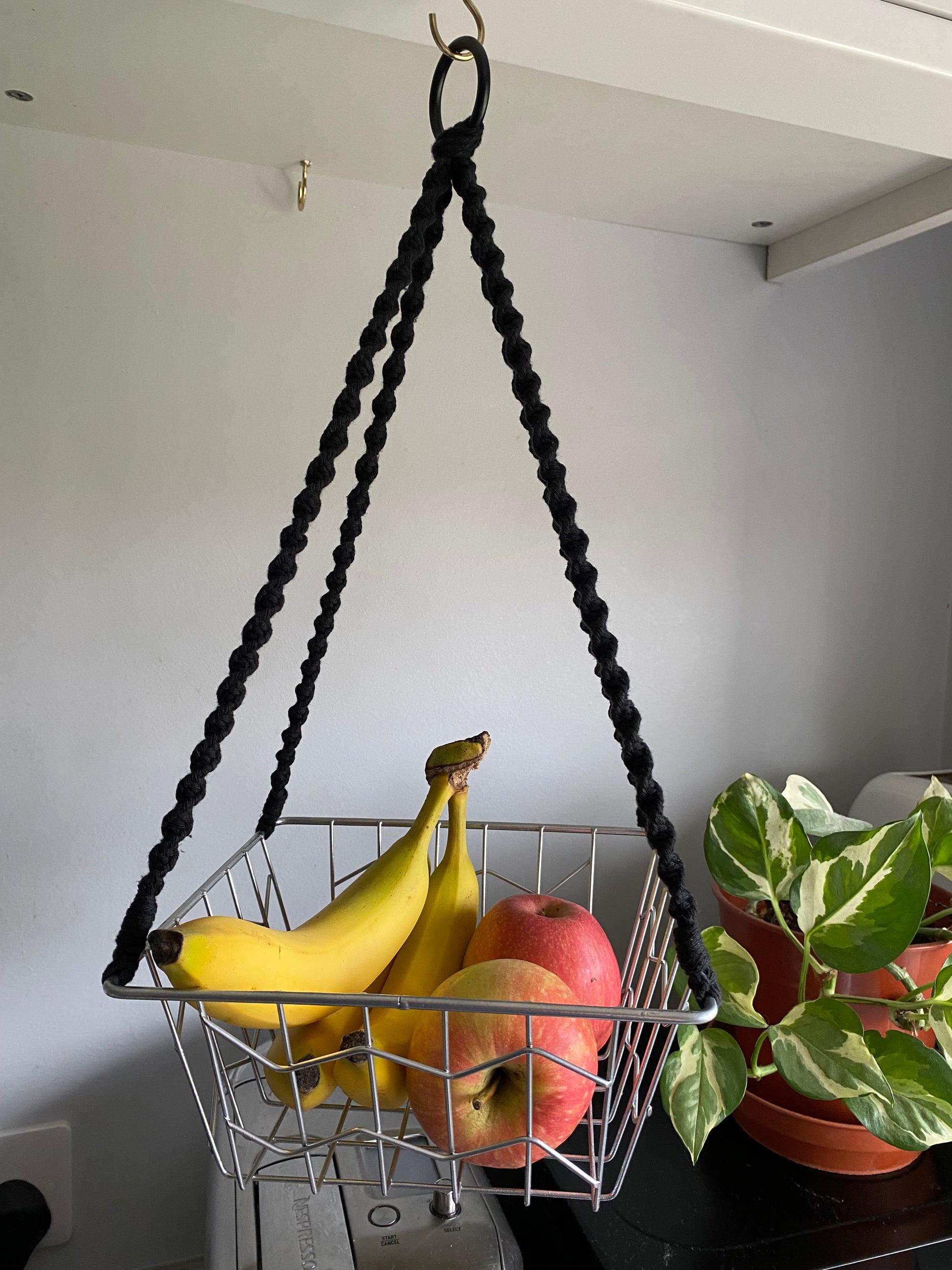 Bananas and apples in a metal basket, suspended by black macrame ropes on a metal hoop, hanging under a white kitchen cabinet next to a variegated houseplant. 