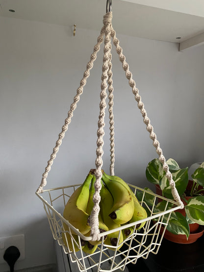 Bananas in a metal basket, suspended by natural macrame ropes on a metal hoop, hanging next to a variegated houseplant. 
