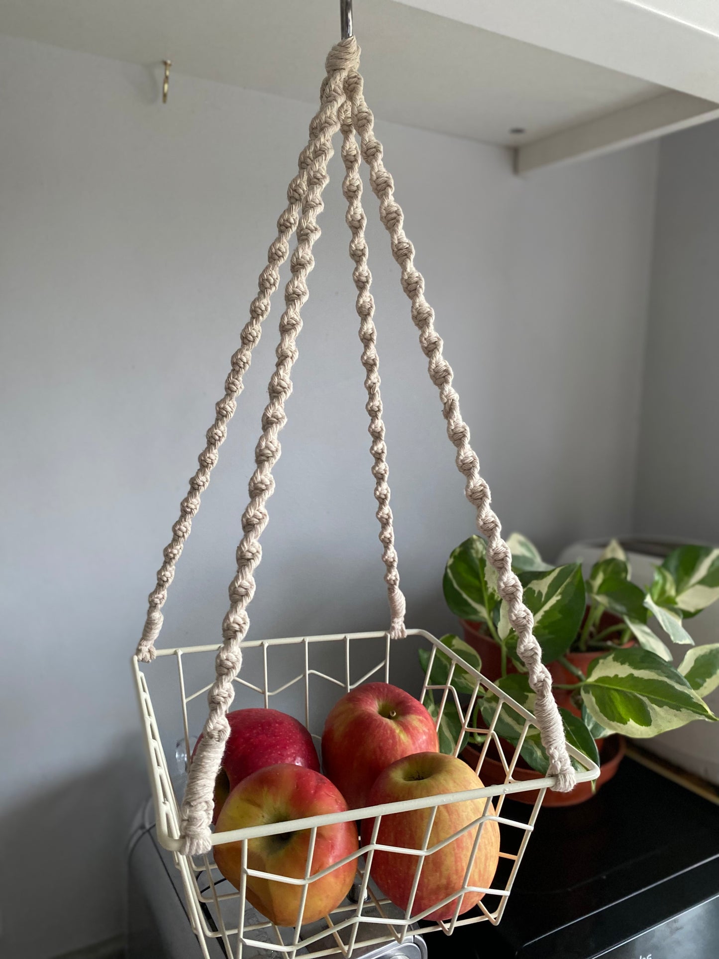 Small apples in a metal basket, suspended by natural macrame ropes on a metal hoop, hanging under a white kitchen cabinet next to a variegated houseplant. 