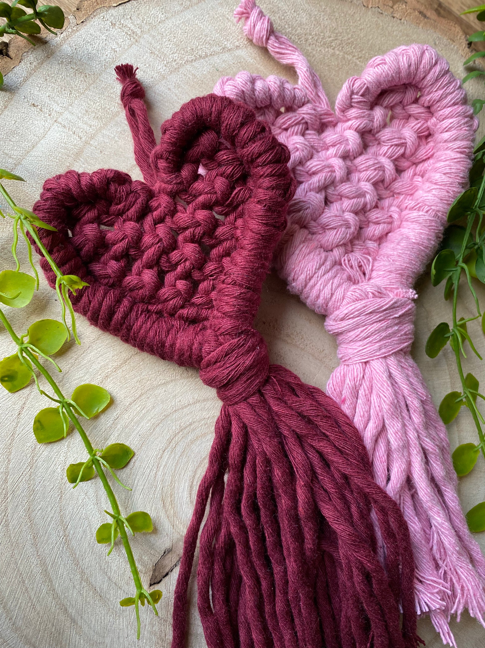 A pair of handmade heart shaped, macrame door hangers laying next to each other on a wooden surface. One is made with a deep red cotton and the other is made with a baby pink cotton. 