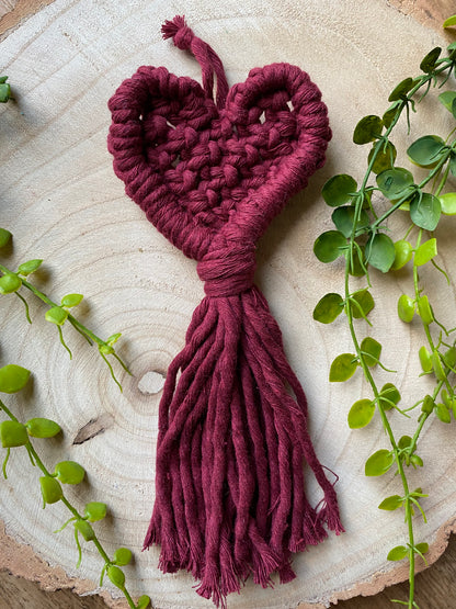 A handmade heart shaped, macrame door hanger on a wooden surface. The hanger is made with a deep red cotton yarn and has a long tassel at the bottom. 