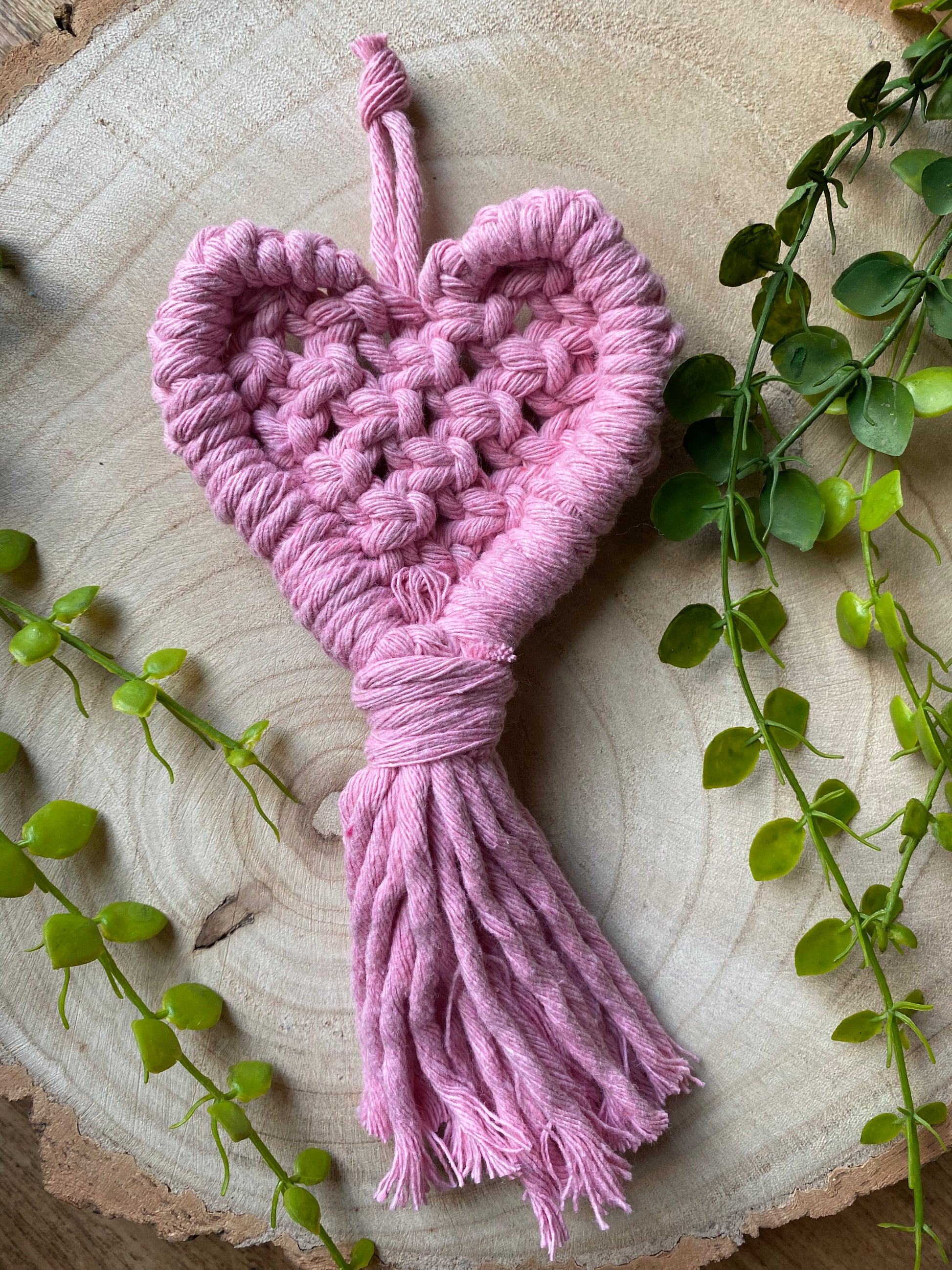 A handmade heart shaped, macrame door hanger laying on a wooden surface. The hanger is made with a baby pink cotton yarn. 