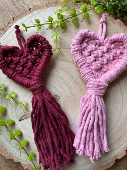 Two handmade heart shaped, macrame door hangers placed on a wooden surface. The hangers are made with deep red and baby pink cotton yarn. 