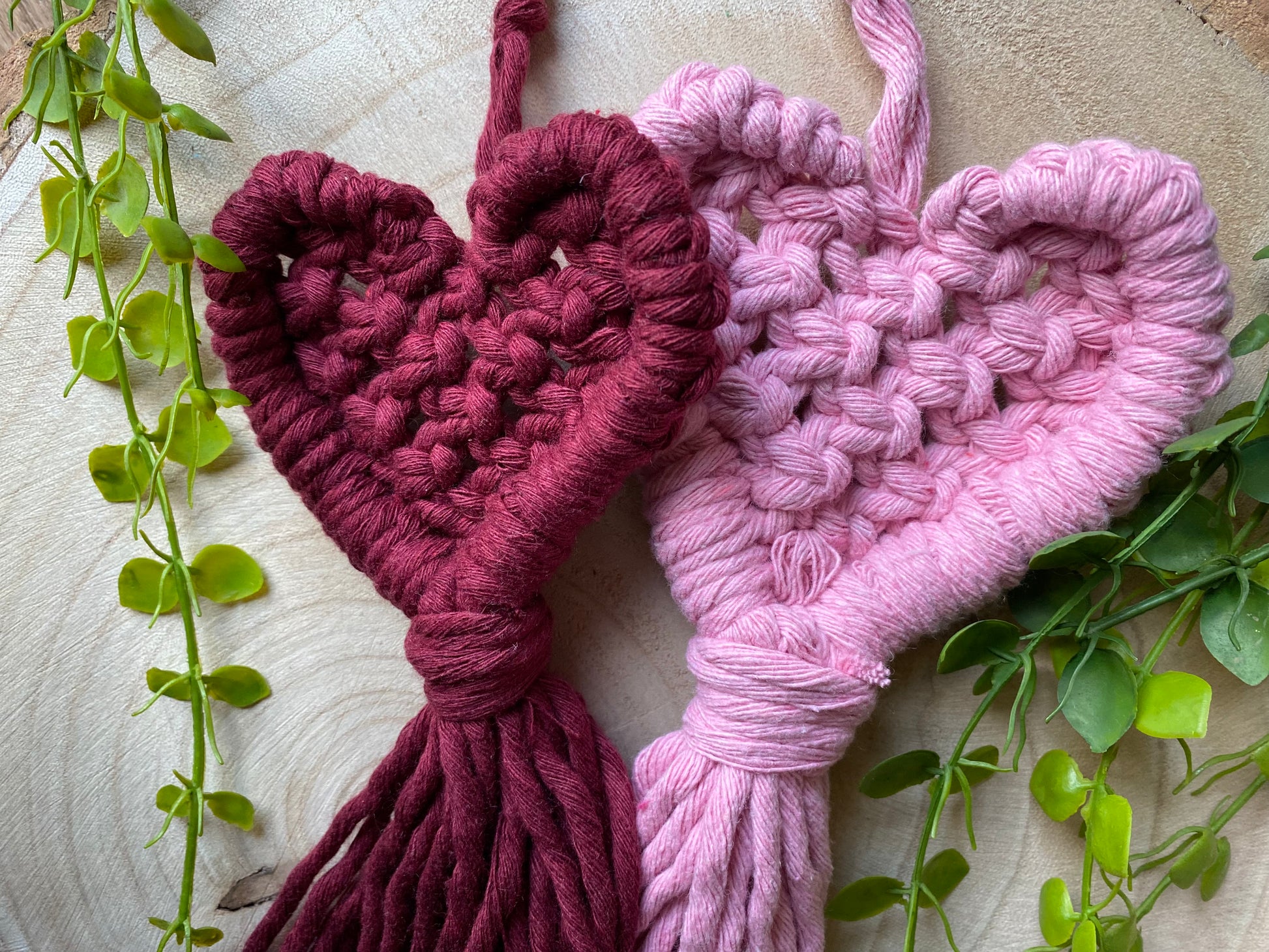 Close up of two handmade heart shaped, macrame door hangers on a wooden surface. The hangers are made with deep red and baby pink cotton yarns. 