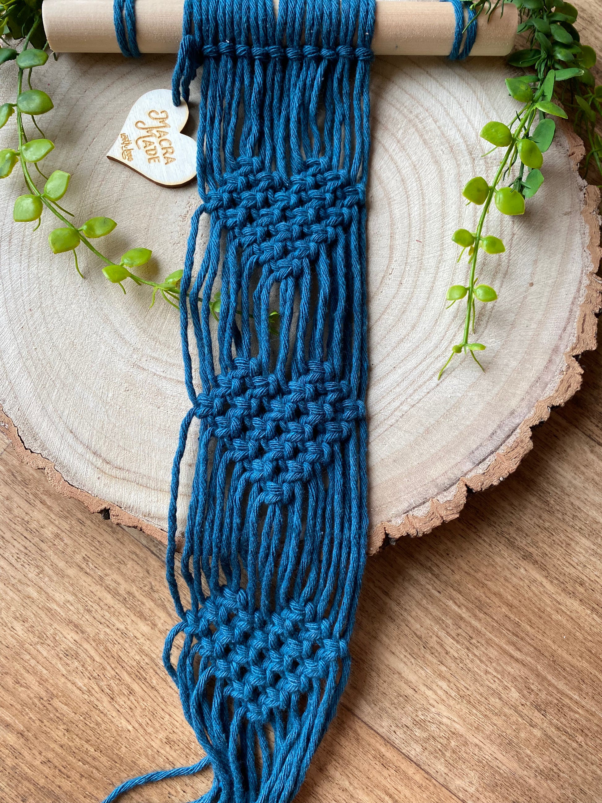 A macrame plant hanger made with petrol blue yarn on a wooden dowel, with knotted heart detail, laid out across a round wooden surface.