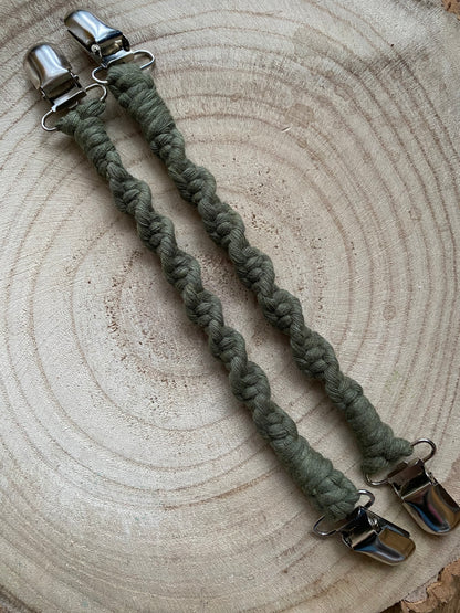 A pair of handmade macrame mitten clips made with avocado green yarn with metal clips on each end, on a wooden board.