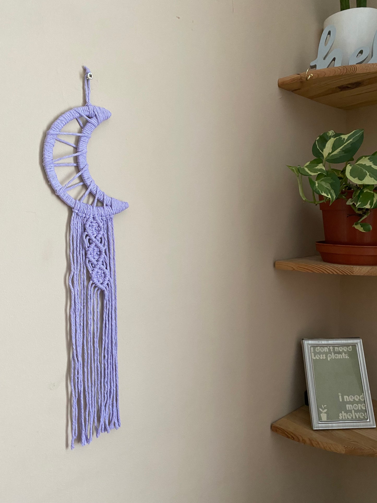 A macrame wall hanging in the shape of a crescent moon, wrapped in lilac purple yarn, with long knotted fringe detail hanging on a beige wall next to a set of wooden corner shelves. 