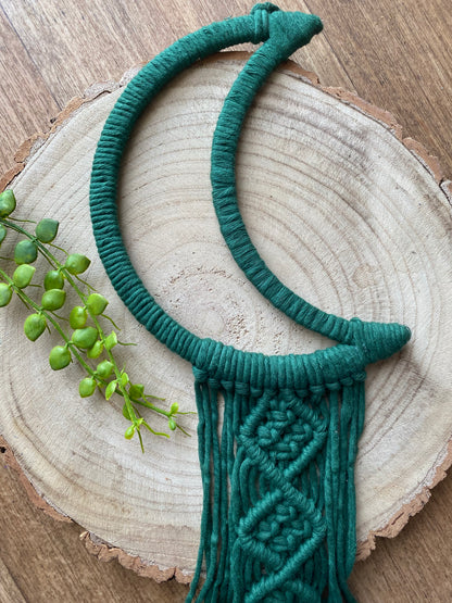 A macrame wall hanging in the shape of a crescent moon, wrapped in pine green yarn, with knotted fringe detail. Laid across a round wooden surface. 