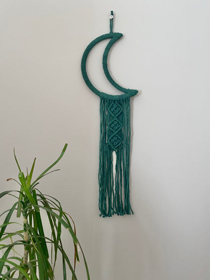 A macrame wall hanging in the shape of a crescent moon, wrapped in pine green yarn, with long knotted fringe detail hanging on a beige wall next to a tall green houseplant. 