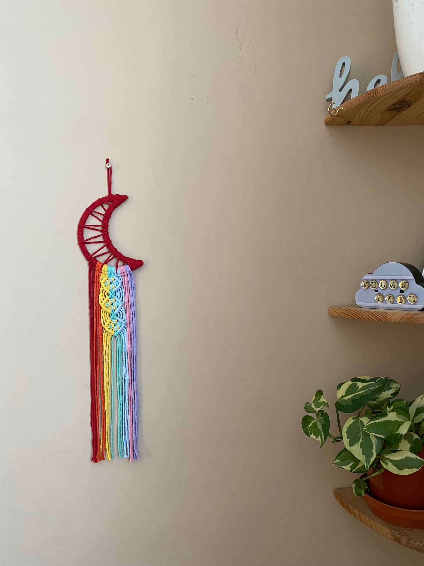 A macrame wall hanging in the shape of a crescent moon, wrapped in red yarn, with long rainbow coloured knotted fringe detail hanging on a beige wall next to a set of wooden corner shelves. 