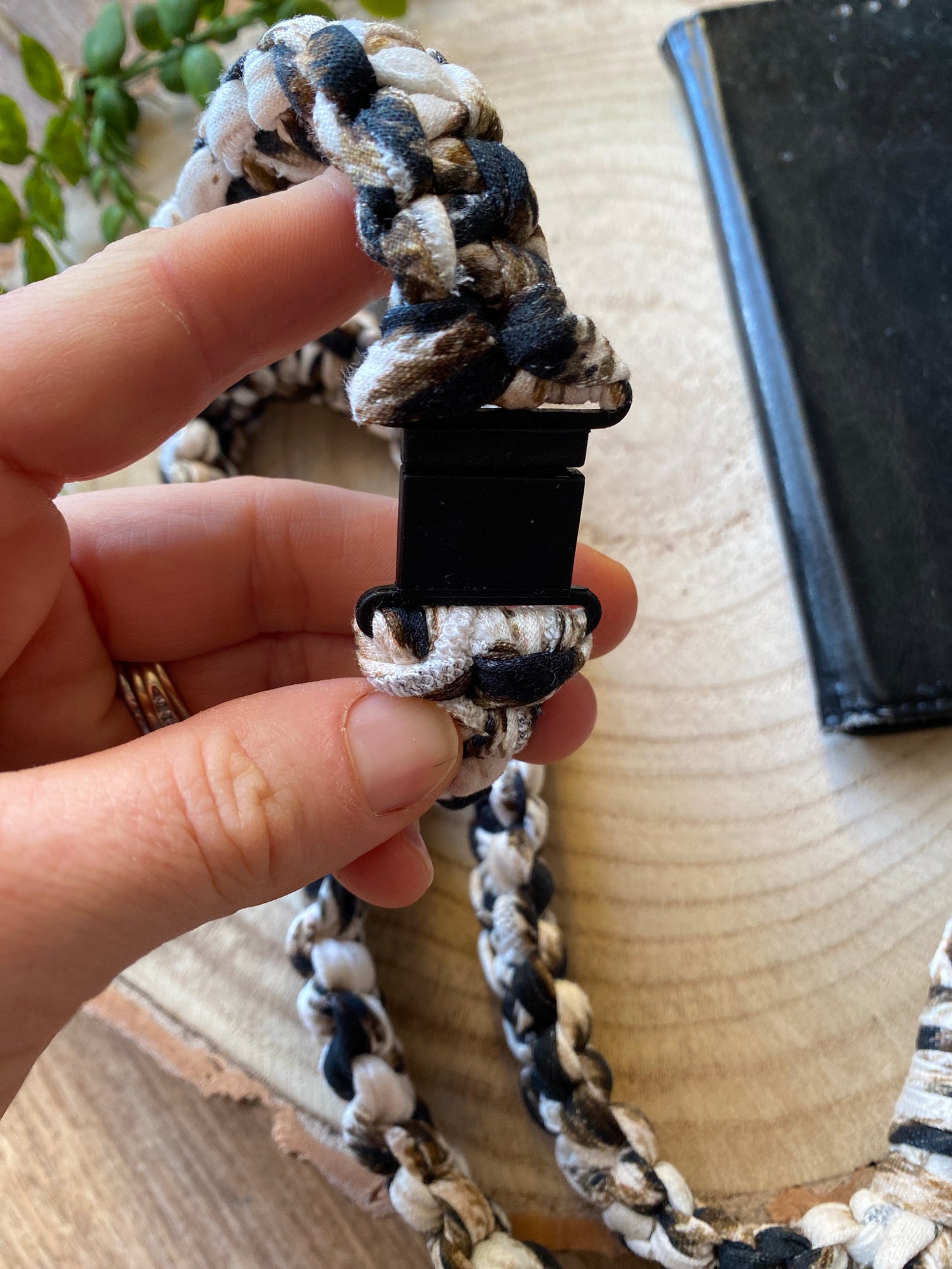 A hand holding a black plastic safety break clasp, which is part of a macrame phone holder lanyard, made with animal print t shirt yarn. 