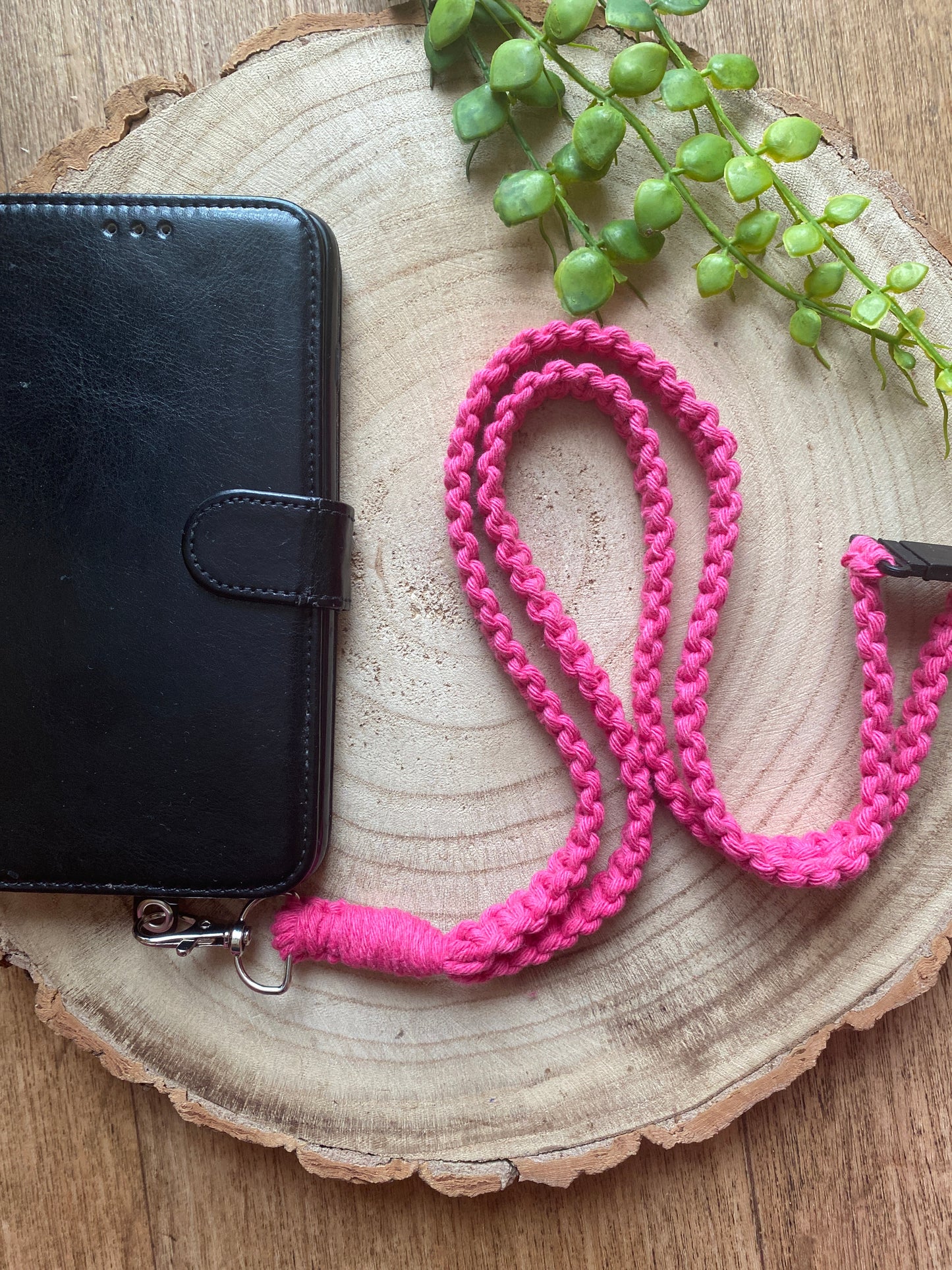A black leather phone case on a wooden board with a macrame phone lanyard made with bright pink yarn attached to it with a metal clasp. 