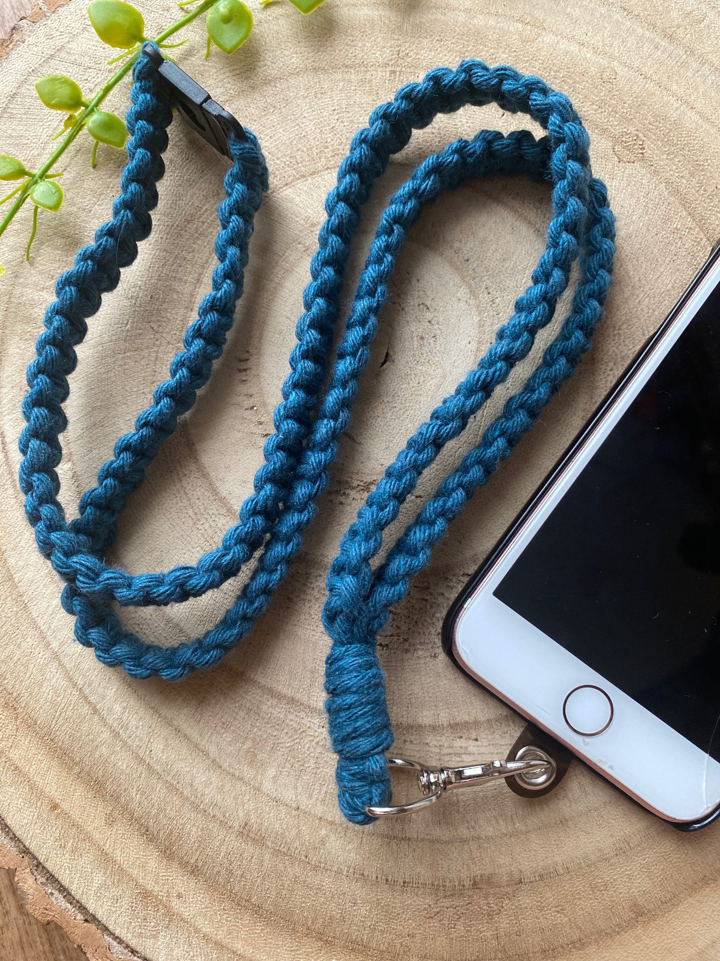An iPhone on a wooden board with a petrol blue macrame phone lanyard attached to it with a metal clasp. 