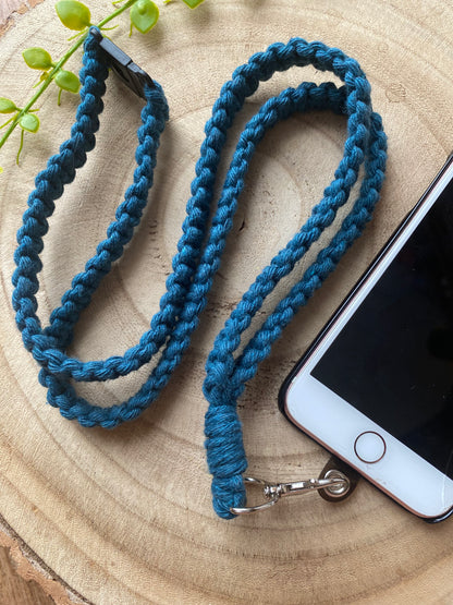An iPhone on a wooden board with a petrol blue macrame phone lanyard attached to it with a metal clasp. 