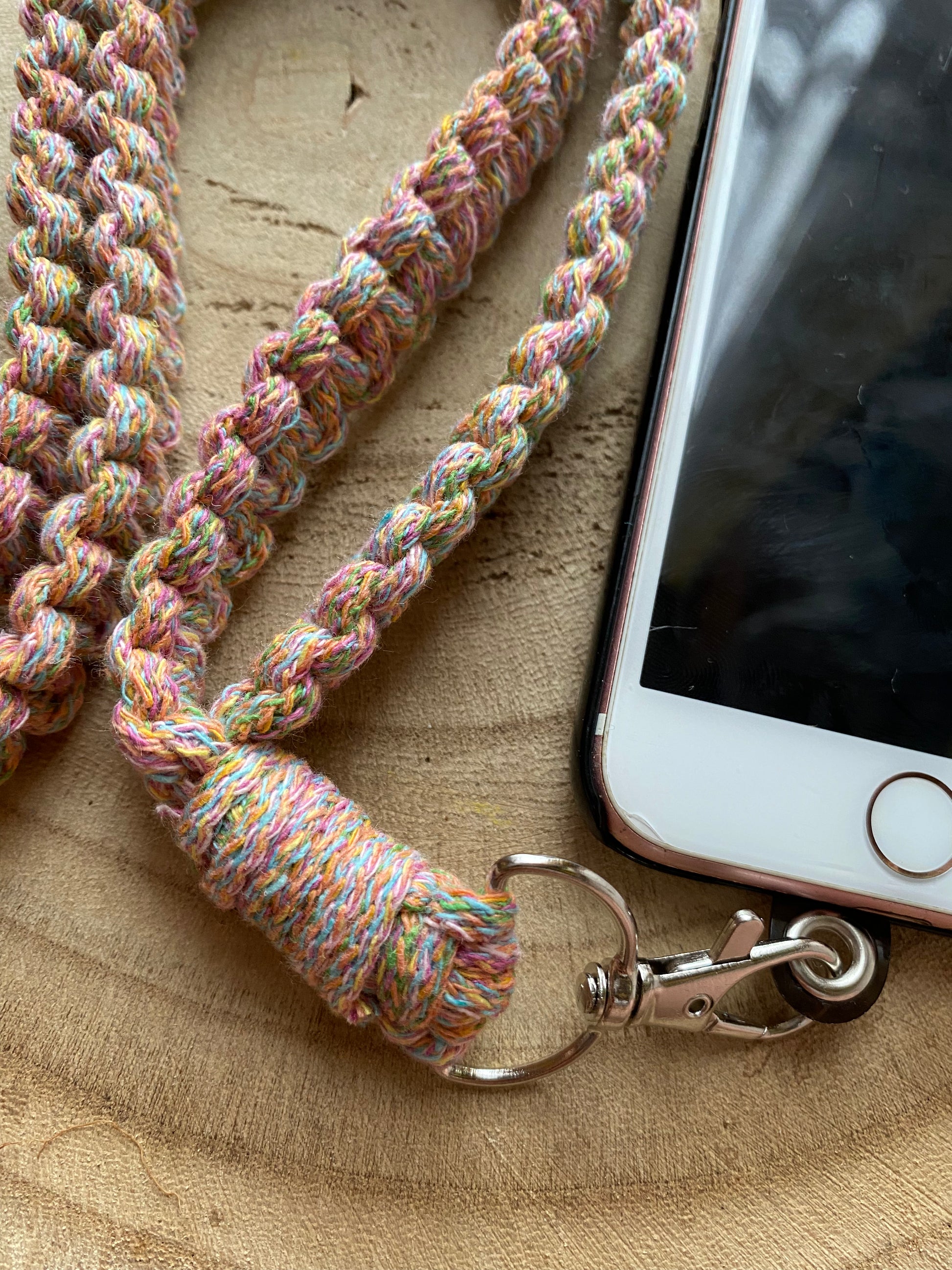 Close up of aniPhone on a wooden board with a rainbow macrame phone lanyard attached to it with a metal clasp. 