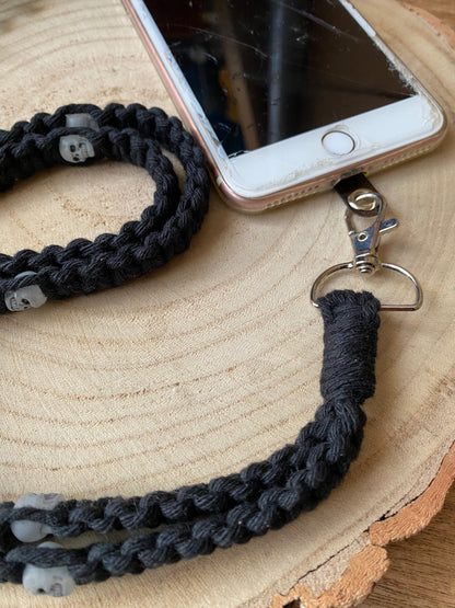 An iPhone on a wooden board with a black macrame phone lanyard with skull beads attached to it with a metal clasp. 
