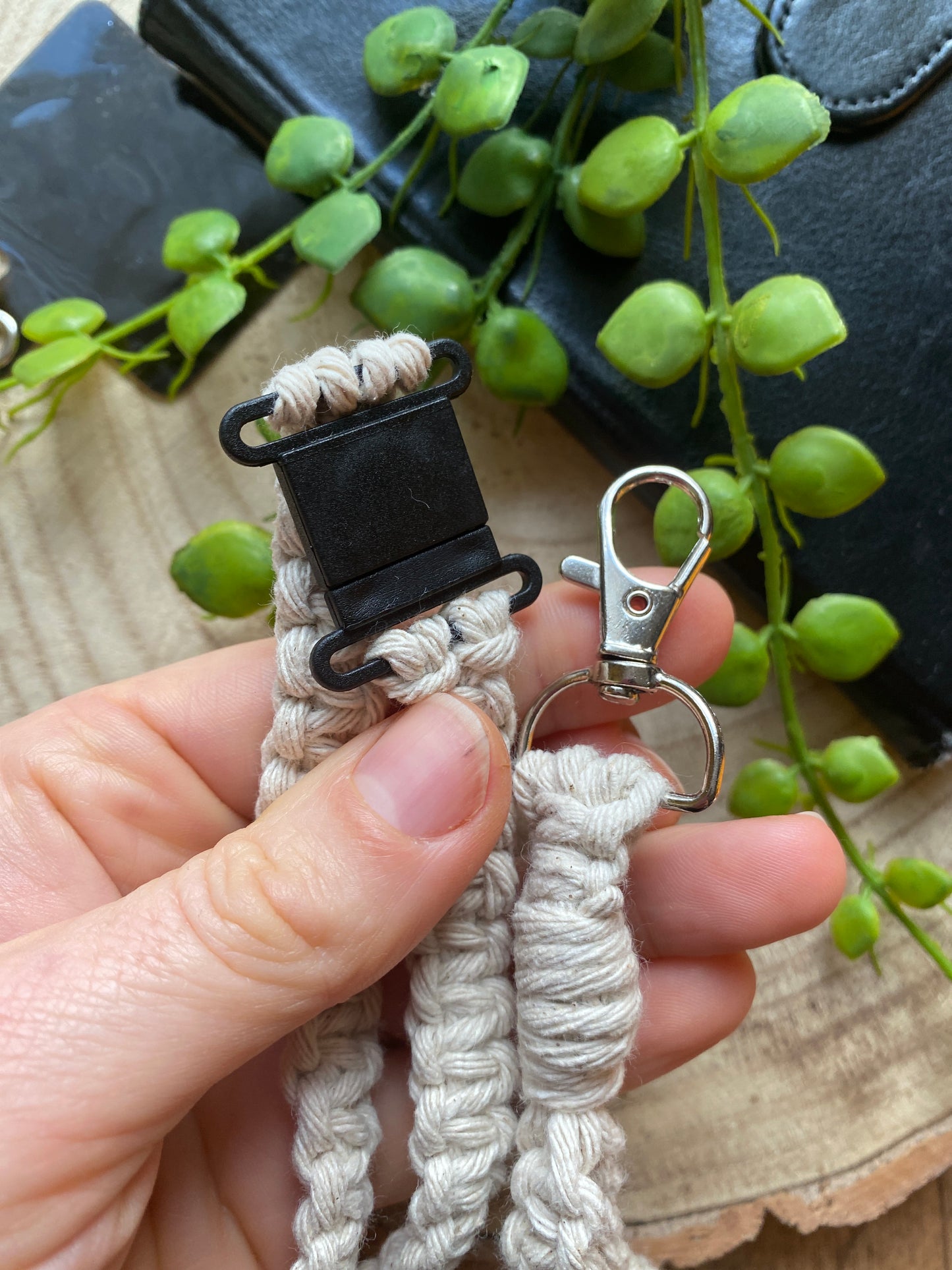 A hand holding a black plastic safety break clasp, and a metal clasp, each being part of a macrame phone holder lanyard made with taupe yarn. 