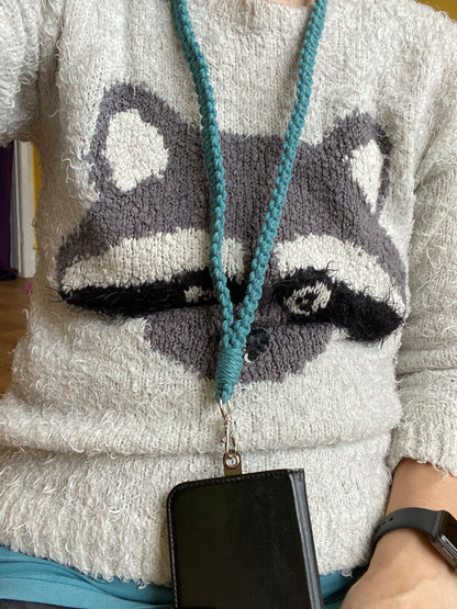 A woman wearing a jumper with a racoon pattern on it, has a teal blue macrame lanyard around her neck, which is attached to a black leather phone case. 