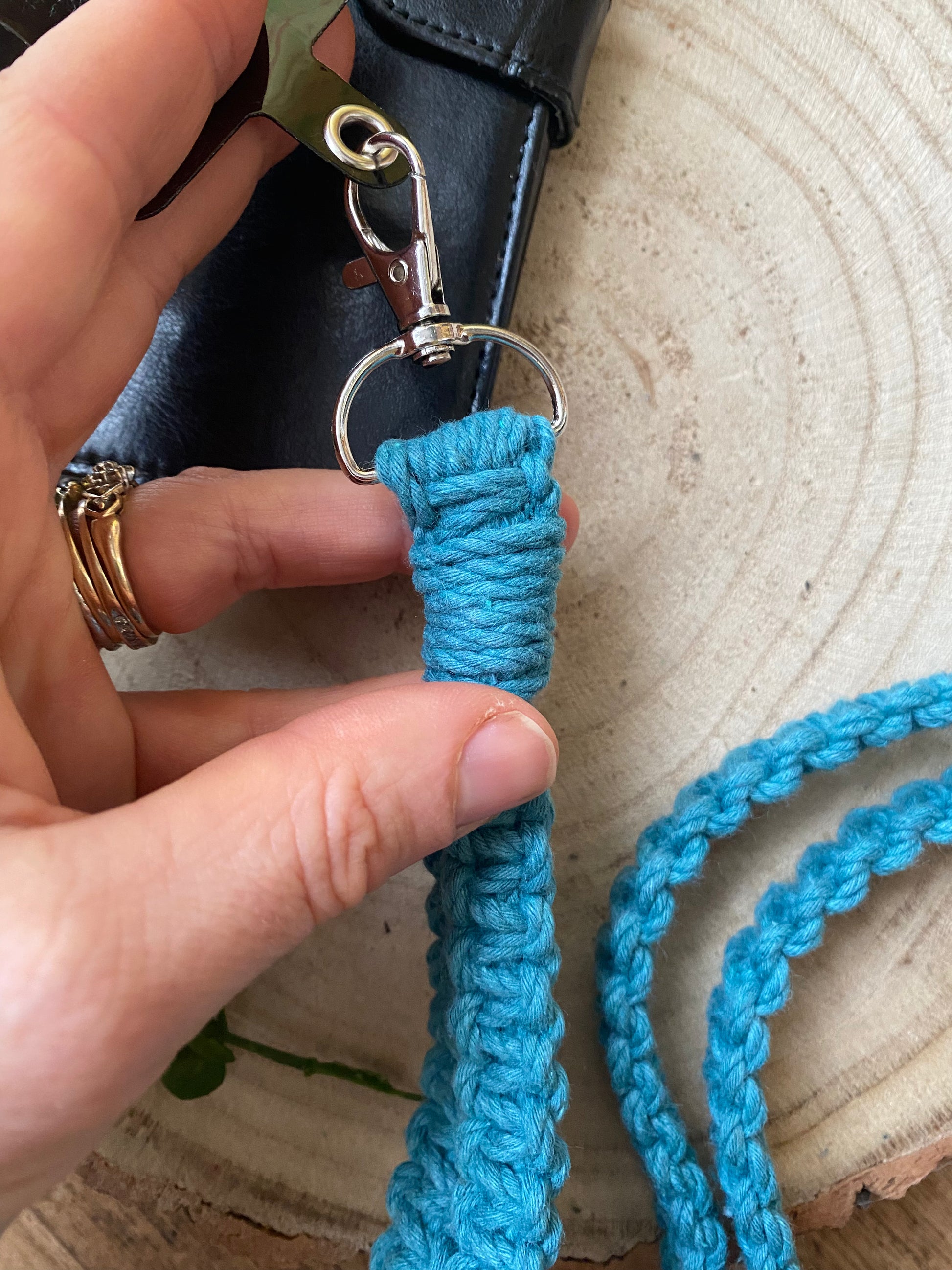 A hand holding the metal clasp on the end of a macrame phone holder lanyard, made with teal blue yarn. 