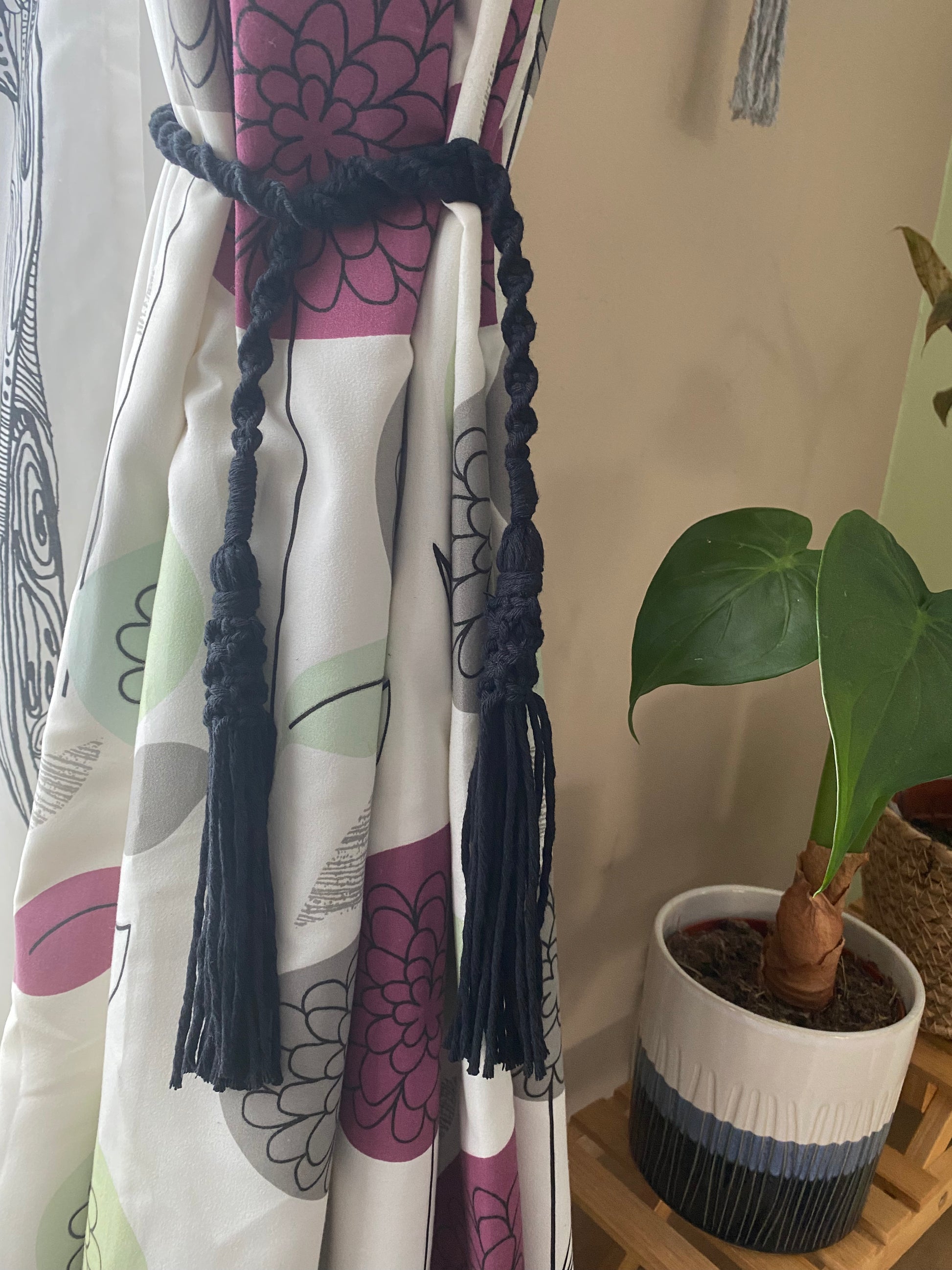 A homely scene featuring patterned curtains held back by black knotted macrame tassel tiebacks, and a green plant in a ceramic pot. 
