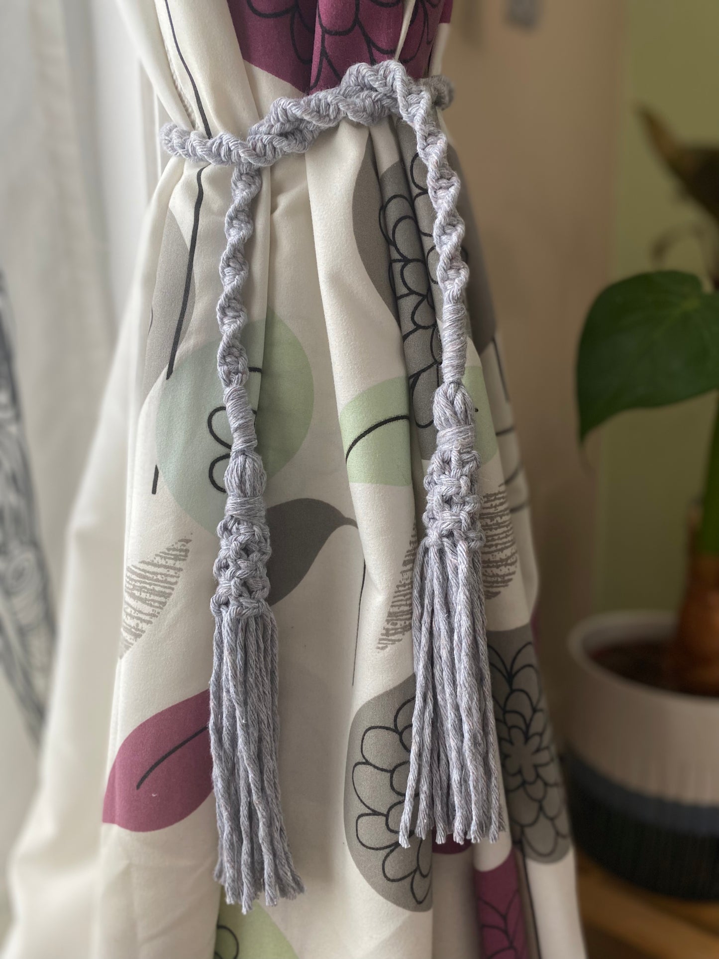 A homely scene featuring patterned curtains held back by knotted macrame tassel tiebacks, made with light grey yarn. 