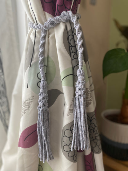 A homely scene featuring patterned curtains held back by knotted macrame tassel tiebacks, made with light grey yarn. 