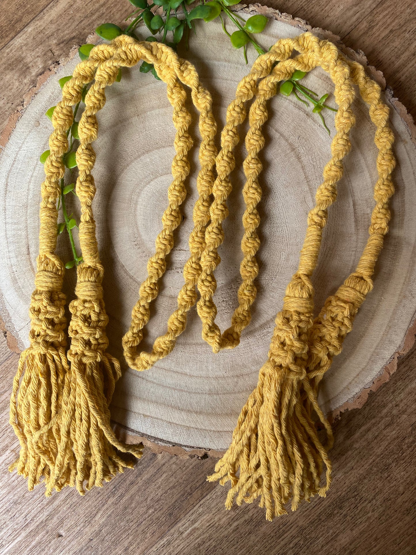 A pair of knotted macrame tassel tiebacks, made with mustard yellow yarn, laid out next to each other on a round wooden board. 