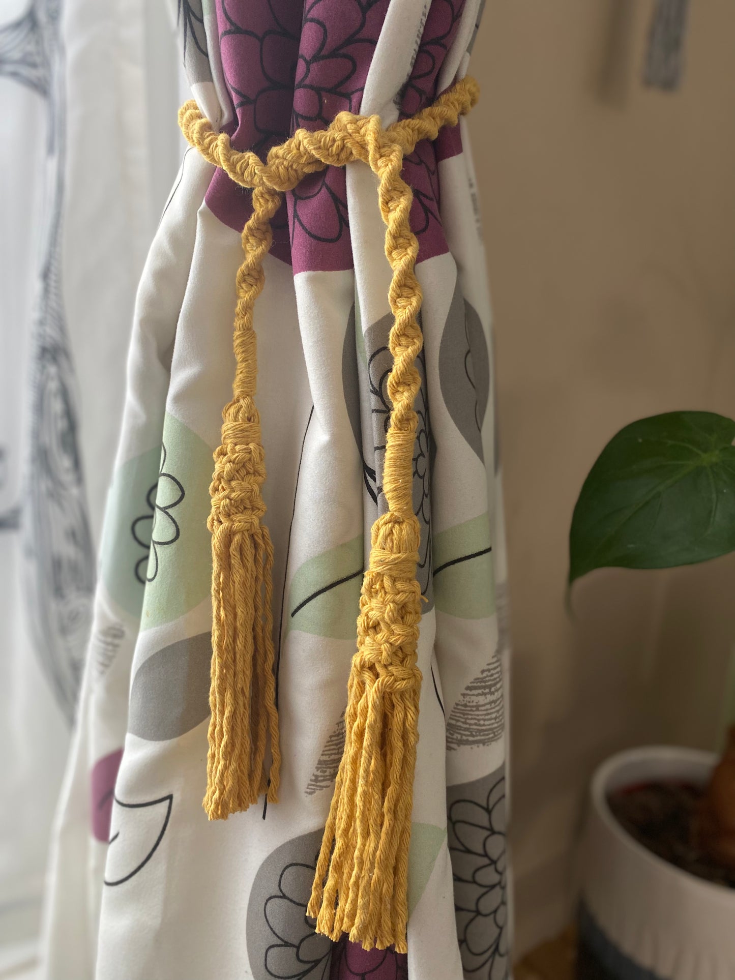 A homely scene featuring patterned curtains held back by knotted macrame tassel tiebacks, made with mustard yellow yarn. 