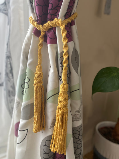 A homely scene featuring patterned curtains held back by knotted macrame tassel tiebacks, made with mustard yellow yarn. 