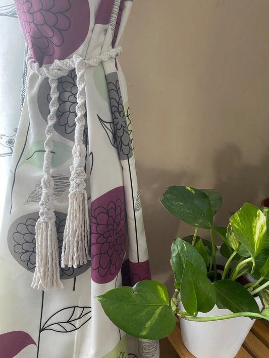 A homely scene featuring patterned curtains held back by knotted macrame tassel tiebacks, and a leafy green plant in a white ceramic pot. 