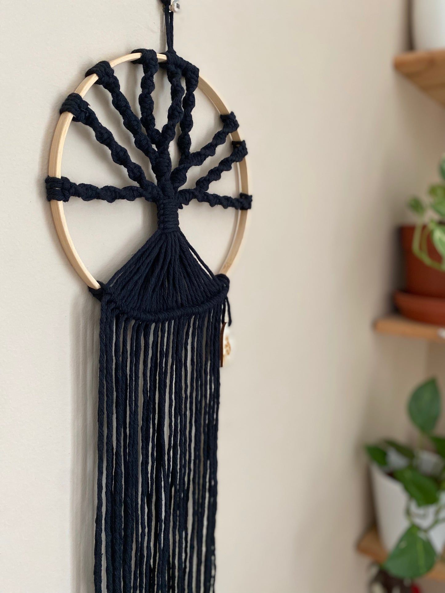 A macrame tree of life wall hanging made on a circular wooden hoop with black knotted yarn, hanging against a beige painted wall. 